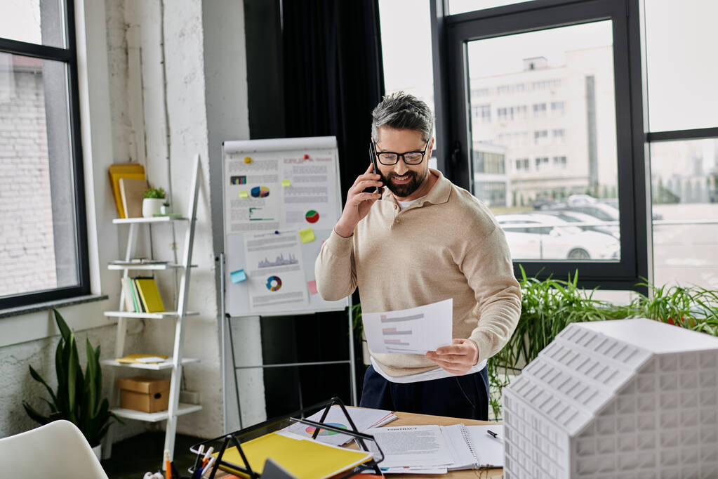Een knappe zakenman met een baard staat in een modern kantoor en bekijkt plannen terwijl hij telefoneert.. - Foto, afbeelding