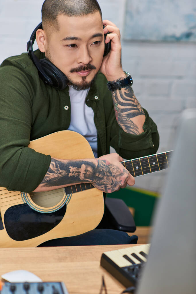 Um homem asiático tatuado em roupas casuais toca uma guitarra acústica enquanto usa fones de ouvido em seu estúdio de gravação em casa. - Foto, Imagem