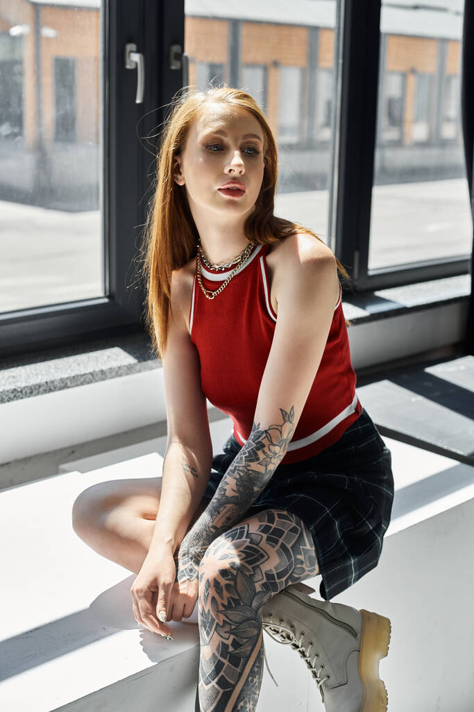 Egy tetovált fiatal nő ül egy fehér párkányon piros felsőt és kockás szoknyát viselve.. - Fotó, kép