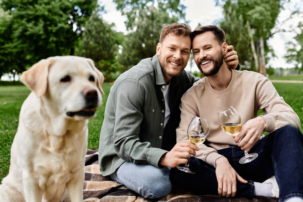 Una pareja gay barbuda disfruta de un picnic con su labrador en un parque verde. Están sonriendo y sosteniendo vasos de vino. - Foto, Imagen