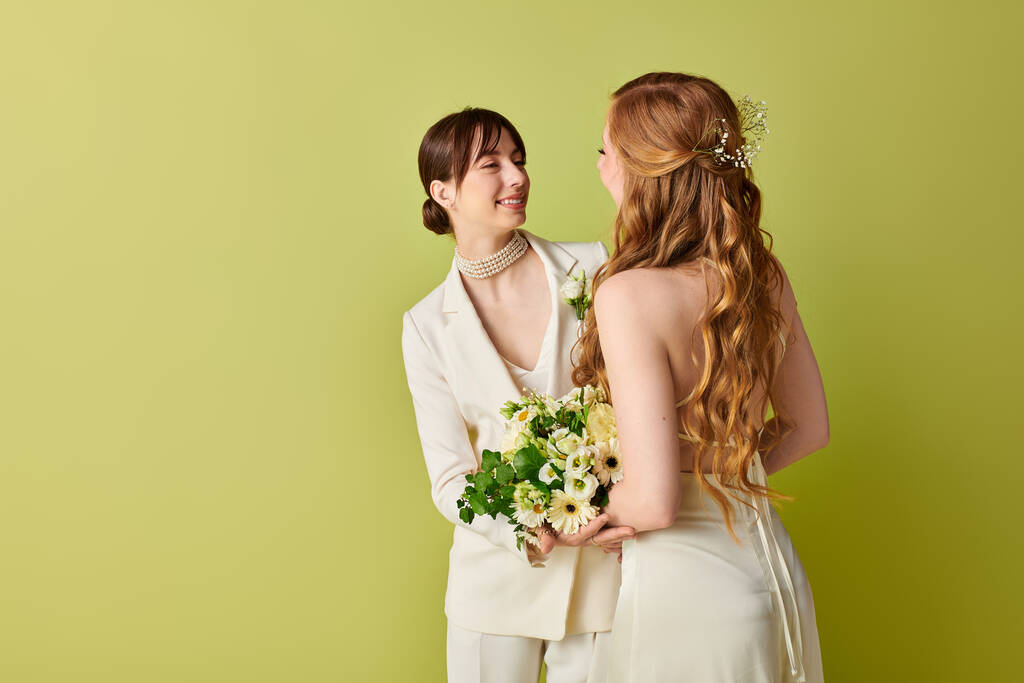 Ein junges lesbisches Paar in weißem Gewand steht bei ihrer Hochzeitszeremonie vor leuchtend grünem Hintergrund in liebevoller Umarmung eng beieinander.. - Foto, Bild