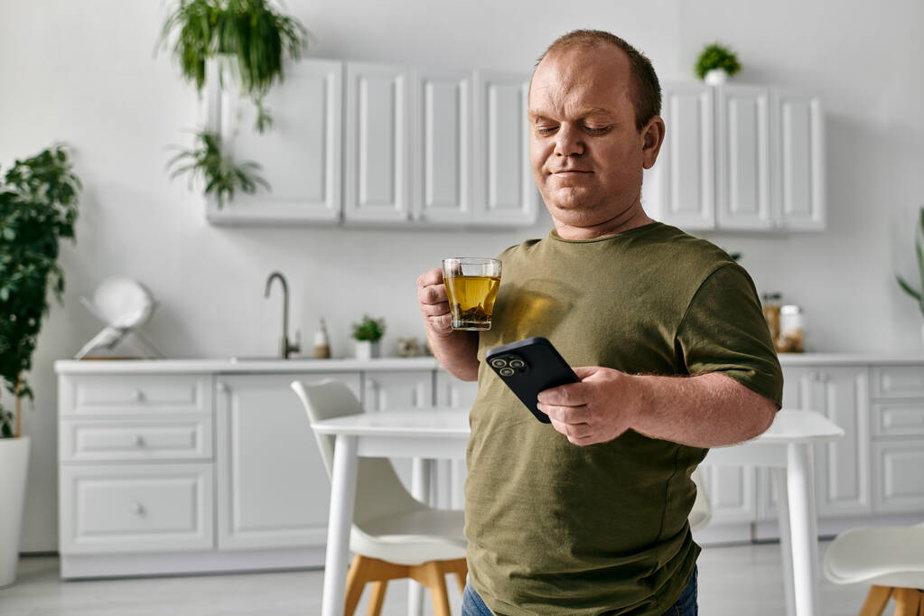 Ένας άντρας με εγκλεισμό απολαμβάνει ένα φλιτζάνι τσάι ενώ τσεκάρει το τηλέφωνό του στην κουζίνα του σπιτιού του.. - Φωτογραφία, εικόνα