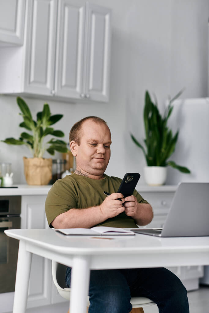 Un uomo con inclusività siede a un tavolo nella sua cucina, vestito con disinvoltura, guardando il suo telefono. - Foto, immagini