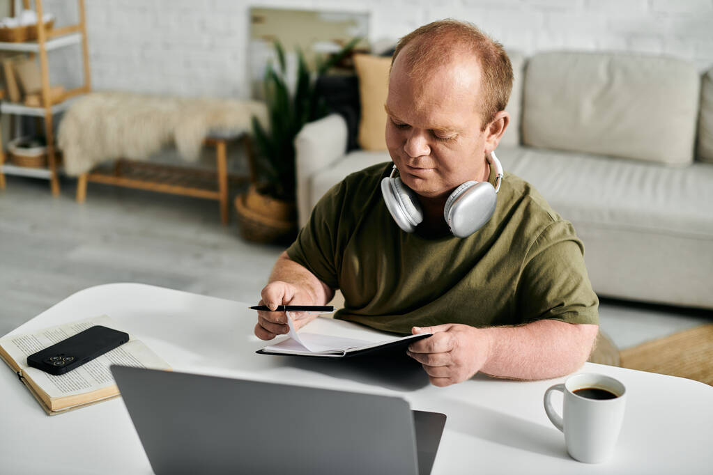 Un homme inclusif s'assoit à une table à la maison, portant des écouteurs et écrivant dans un cahier. - Photo, image