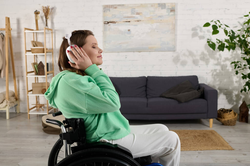 Een jonge vrouw met bruin haar zit in een rolstoel in haar woonkamer naar muziek te luisteren via een koptelefoon.. - Foto, afbeelding