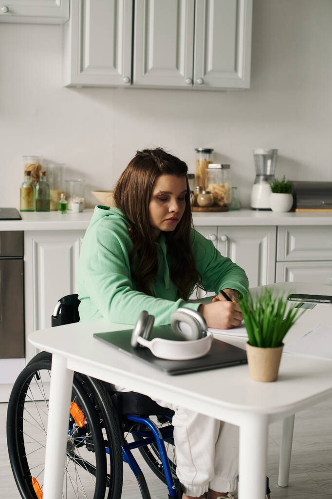 車椅子に乗っている若い女性が自宅のテーブルで働いている. 彼女は緑色のスウェットシャツを着ていて,紙に書いている.. - 写真・画像