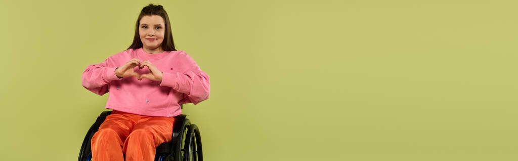 Une jeune femme aux cheveux bruns est assise dans un fauteuil roulant dans un studio, souriante et en forme de cœur avec ses mains. - Photo, image