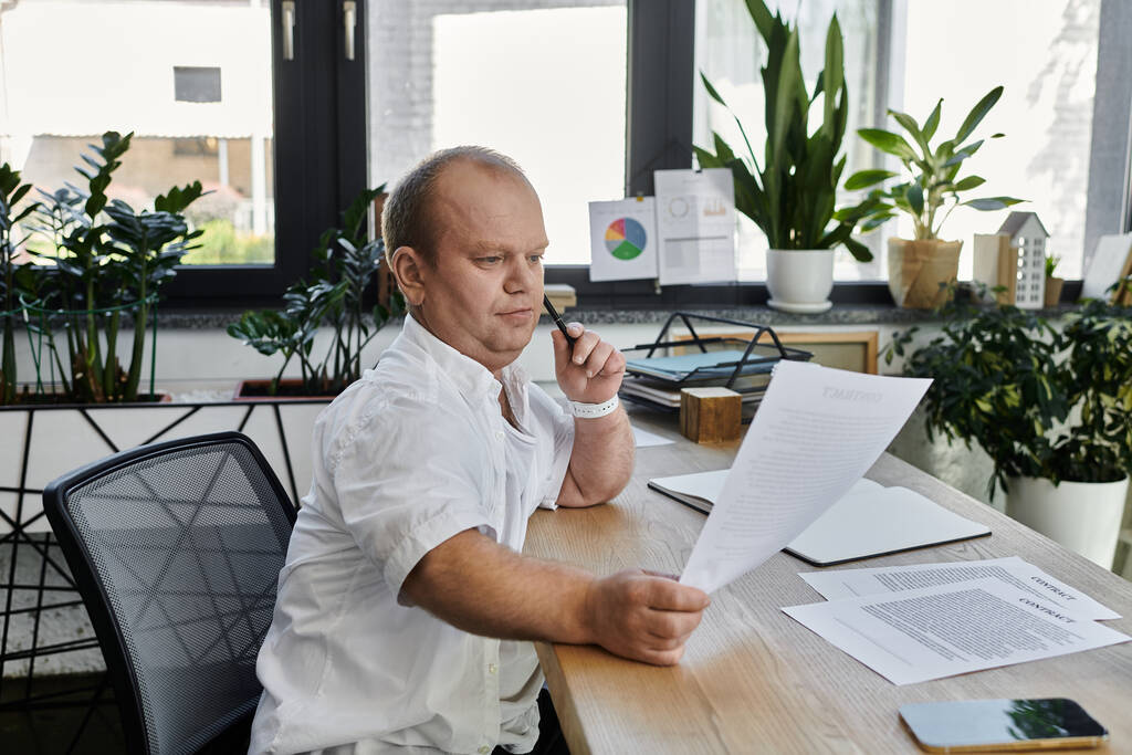 Человек с инклюзивностью сидит за столом в современном офисе, просматривает документы. - Фото, изображение