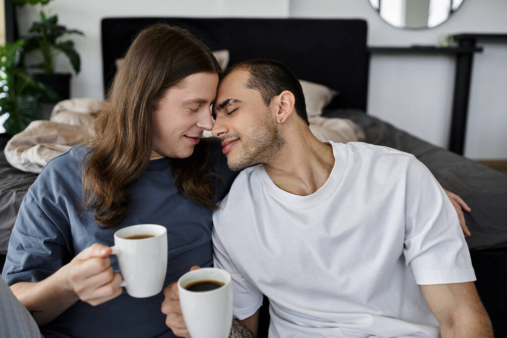 Una joven pareja gay disfruta de una tranquila mañana juntos en su dormitorio, compartiendo una taza de café y un momento tierno. - Foto, imagen
