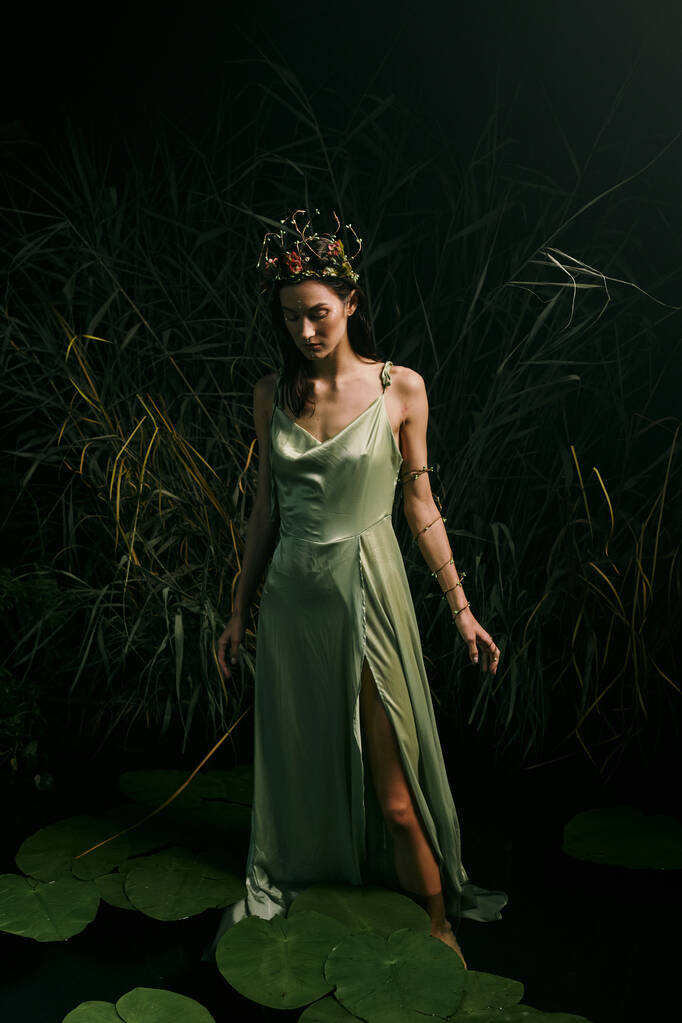 Una mujer está de pie con gracia en las almohadillas de lirio en un pantano, con un vestido verde que fluye y una corona floral. - Foto, imagen