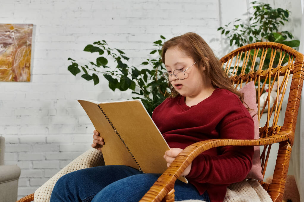 Down sendromlu küçük bir kız evde hasır bir sandalyede oturuyor, kendini bir kitaba kaptırmış.. - Fotoğraf, Görsel