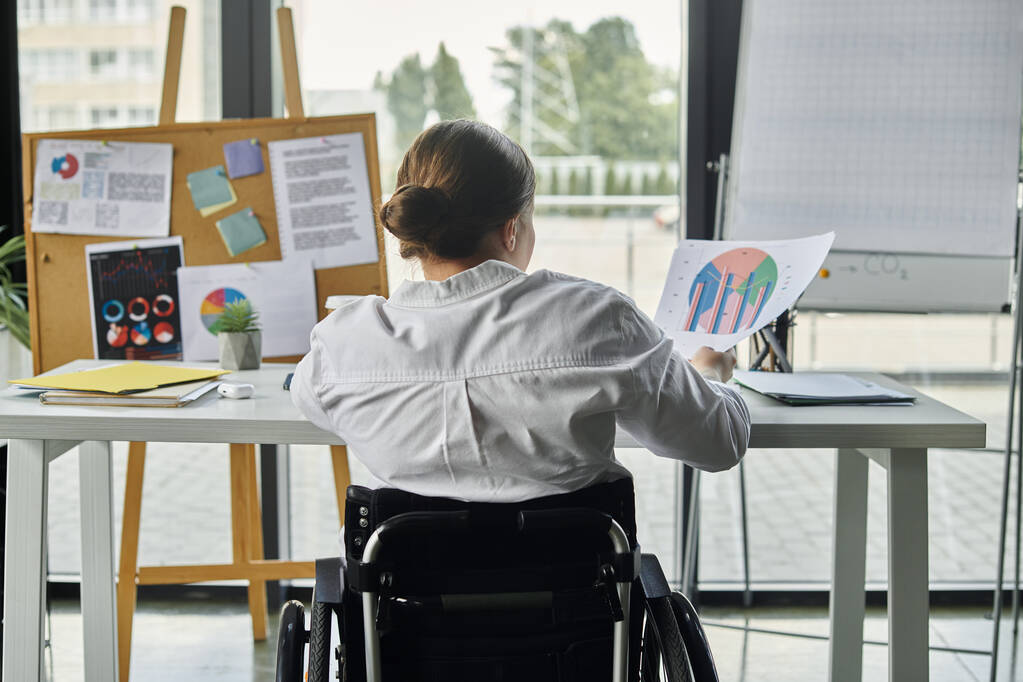 Mladá podnikatelka na invalidním vozíku pracuje za stolem v moderní kanceláři, analyzuje data a přispívá k úspěchu svých týmů. - Fotografie, Obrázek