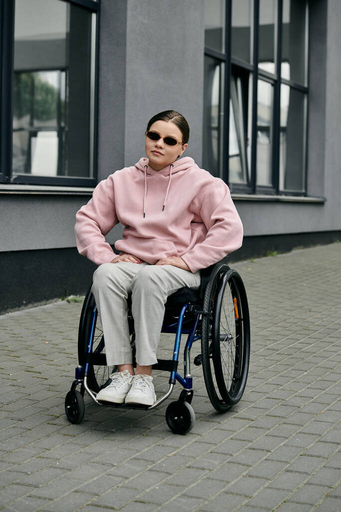 Genç bir kadın bir binanın önündeki kaldırımda tekerlekli sandalyede oturuyor. Pembe bir kapüşonlu ve güneş gözlüğü takıyor.. - Fotoğraf, Görsel