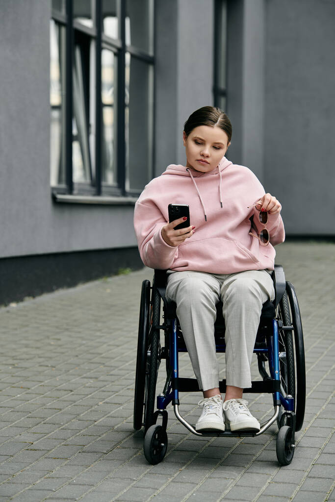 Een jonge vrouw in een roze hoodie zit in een rolstoel op een verhard pad terwijl ze naar haar telefoon kijkt. - Foto, afbeelding