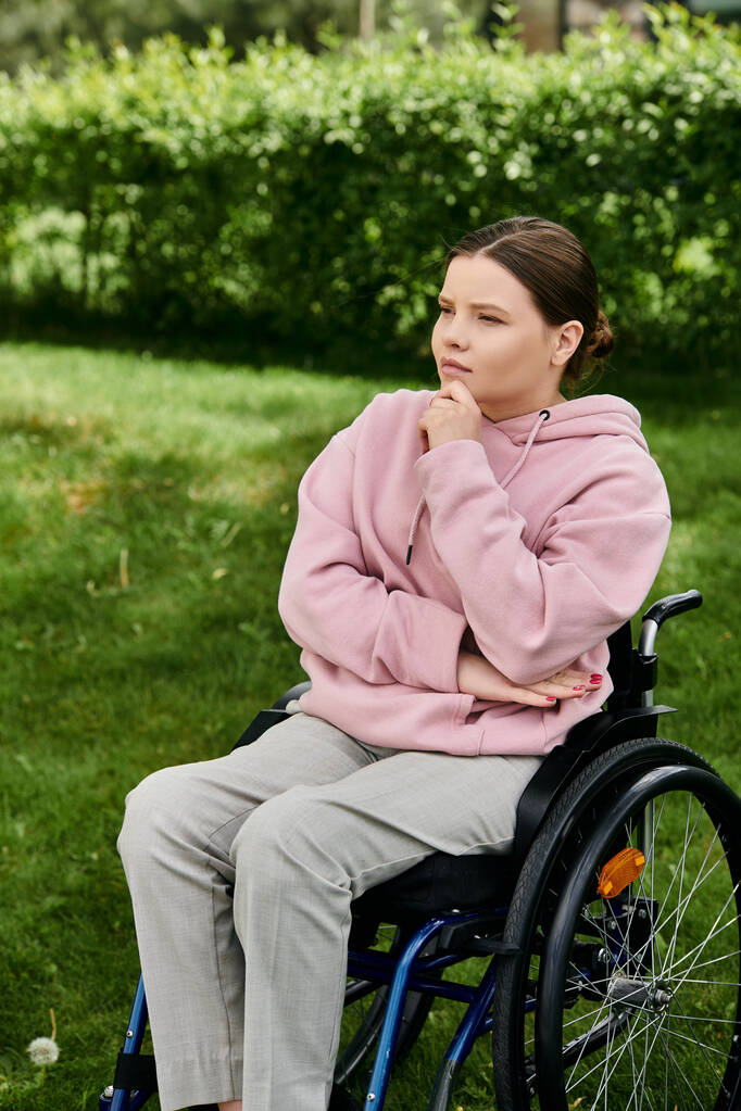 Una joven en una sudadera rosa se sienta pensativamente en una silla de ruedas al aire libre. - Foto, Imagen