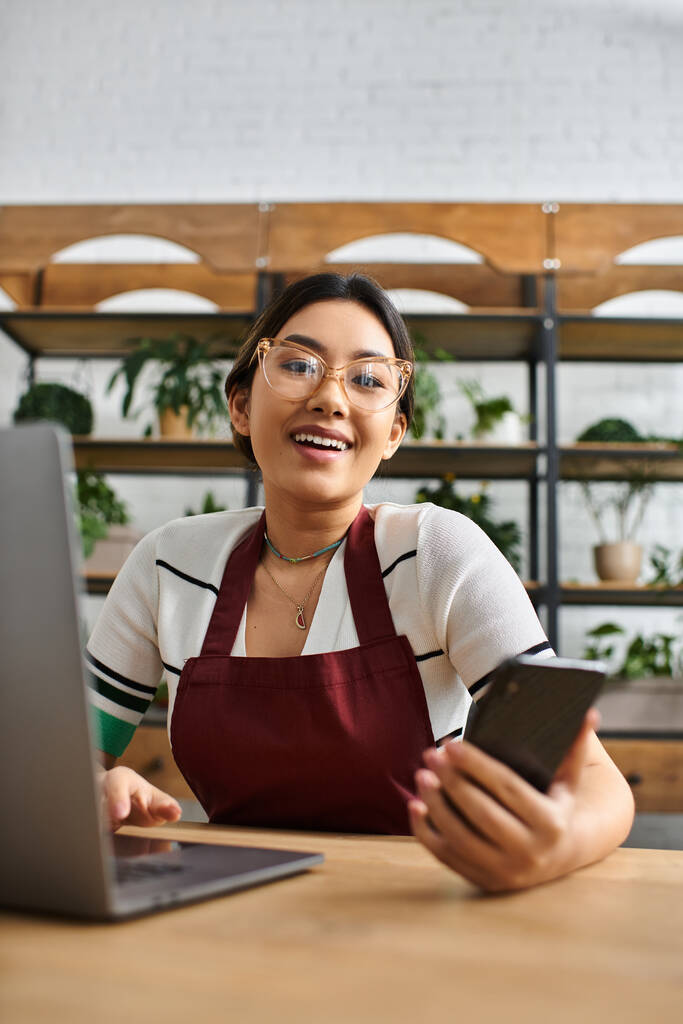 Uma mulher asiática em um avental sorri enquanto trabalhava em um laptop em sua loja de plantas. - Foto, Imagem