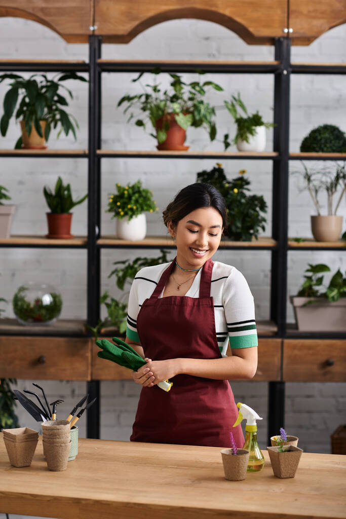 Una joven vistiendo un delantal rojo sonríe mientras trabaja en su tienda de plantas, rodeada de exuberante vegetación. - Foto, Imagen
