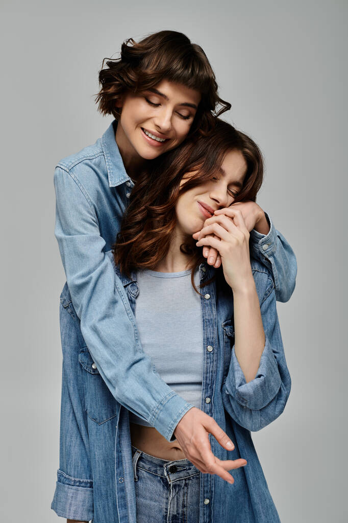 Dwie młode kobiety w dżinsowych ciuchach stawiają czule na szarym tle, pokazując swoje stylowe osobowości i miłość. - Zdjęcie, obraz