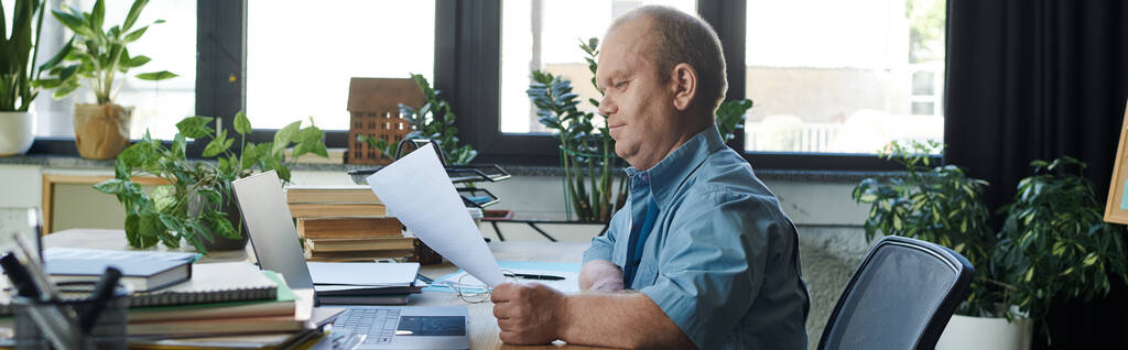 Muž s inkluzivitou sedí za stolem, prohlíží si papíry a pracuje na laptopu. - Fotografie, Obrázek