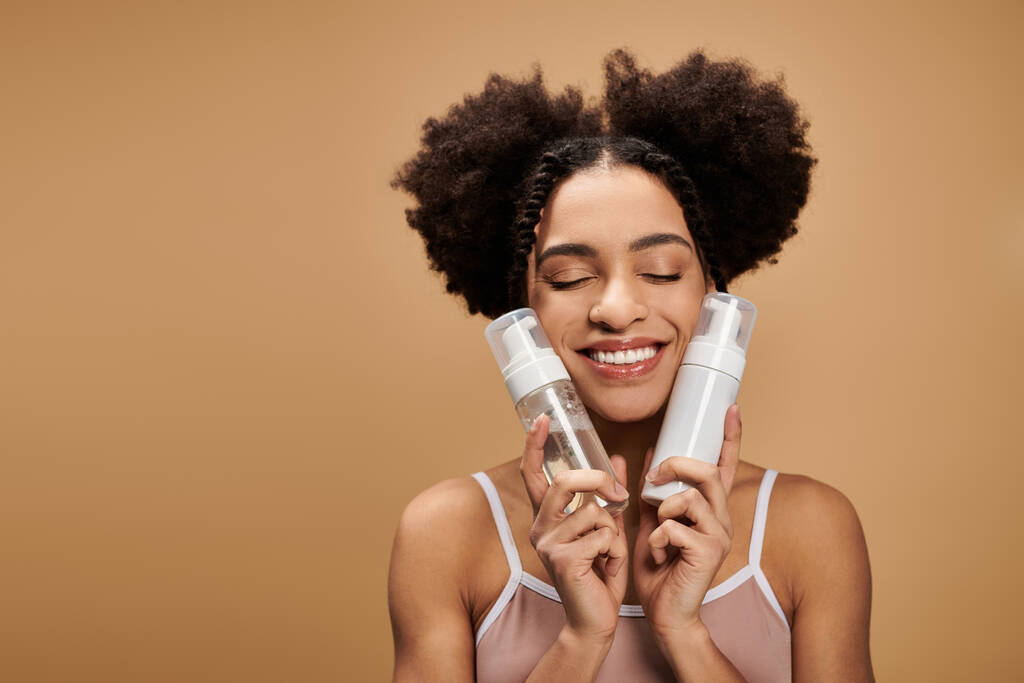 Una giovane donna afroamericana con un bel sorriso tiene due bottiglie di prodotti per la cura della pelle contro il viso. - Foto, immagini
