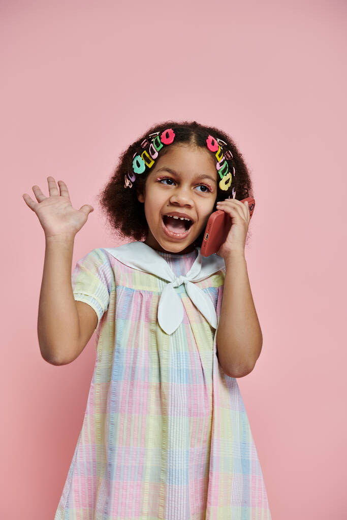 Ένα χαριτωμένο κοριτσάκι με πολύχρωμα κλιπ μαλλιών στέκεται σε ένα πολύχρωμο φόρεμα, χαμογελώντας έντονα, ενώ μιλάμε στο τηλέφωνο. - Φωτογραφία, εικόνα