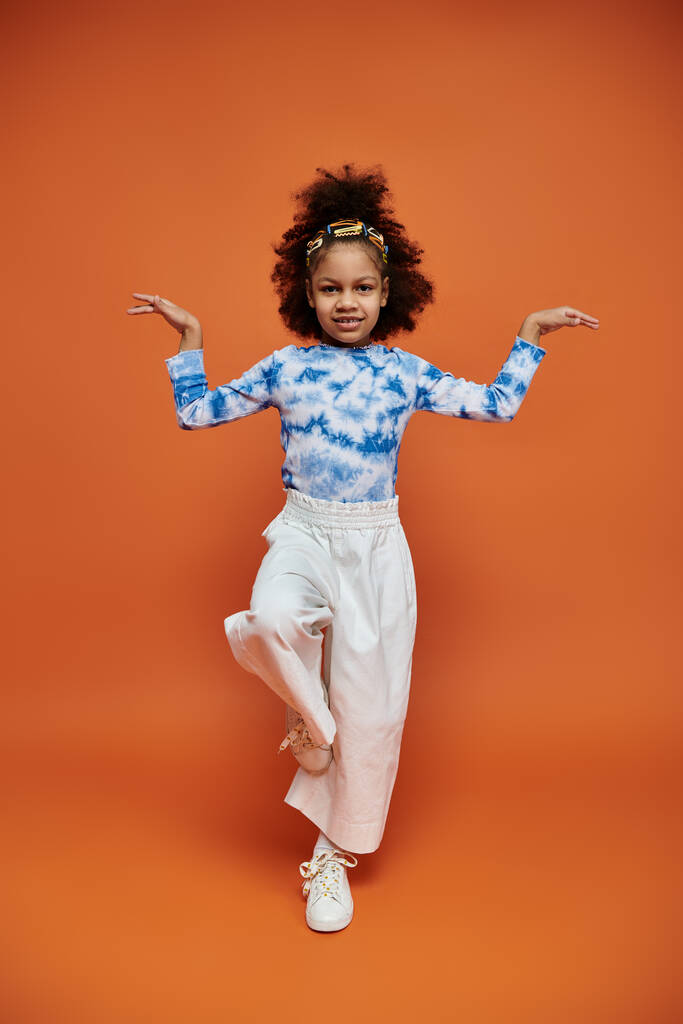 Une jeune afro-américaine avec une coiffure et une tenue tendance pose les bras devant une toile de fond orange. - Photo, image