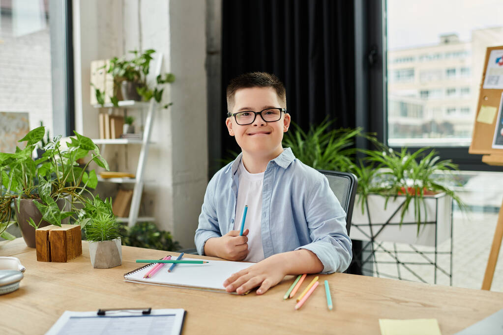 Egy Down-szindrómás kisfiú ül egy íróasztalnál egy modern irodában, mosolyog és rajzol színes ceruzákkal.. - Fotó, kép
