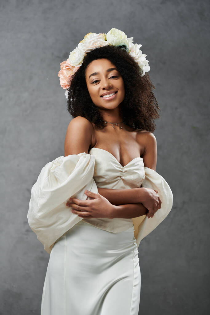 Une belle mariée afro-américaine se tient dans une robe de mariée blanche avec une couronne florale sur un fond gris. - Photo, image