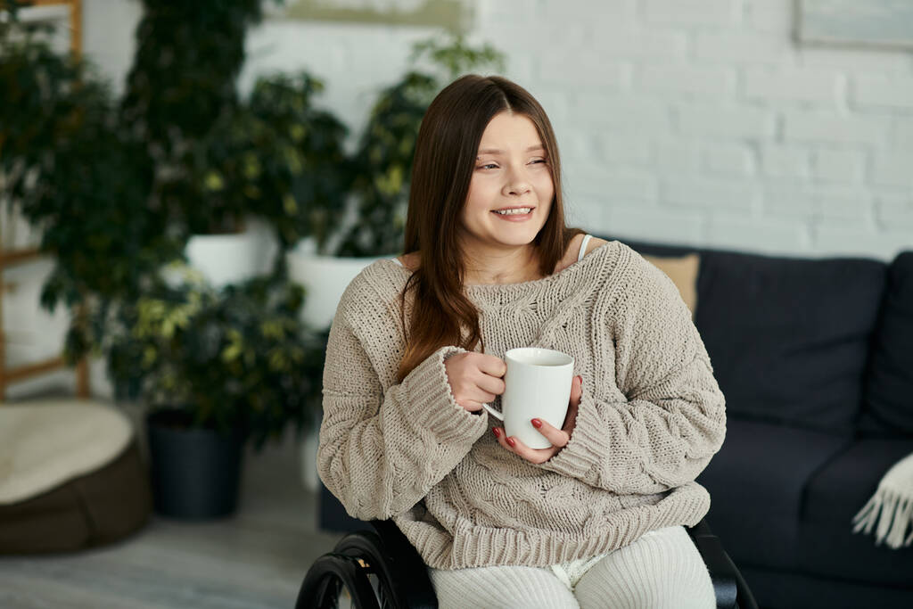 A young woman in a wheelchair sits at home, smiling and holding a mug of coffee. - Photo, Image