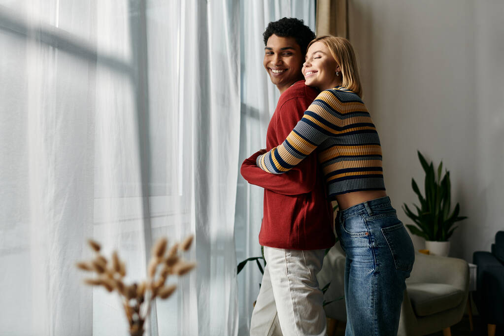 Egy boldog, multikulturális pár ölelkezik egy ablaknál a modern lakásukban.. - Fotó, kép
