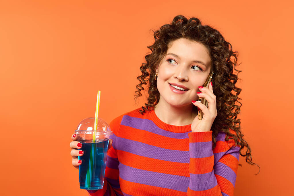 Una mujer joven con el pelo rizado en un vibrante suéter a rayas naranja y púrpura habla en su teléfono mientras sostiene una bebida azul con una paja. - Foto, Imagen