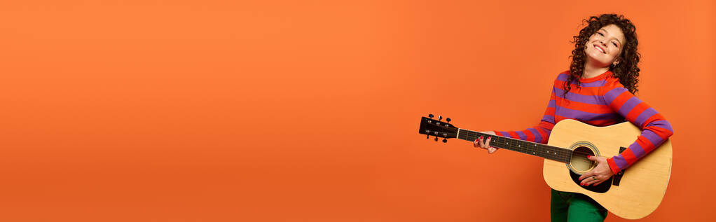 Egy fiatal, göndör hajú nő gitárral pózol, fényesen mosolyogva a narancssárga háttér felé.. - Fotó, kép