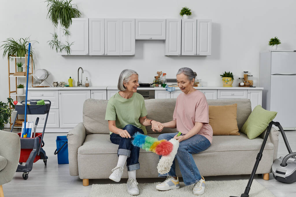 Een volwassen lesbisch paar geniet van het schoonmaken van hun moderne appartement samen. - Foto, afbeelding