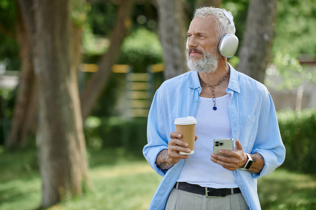 Ein reifer Mann mit Tätowierungen und grauem Bart genießt eine Tasse Kaffee und Musik - Foto, Bild