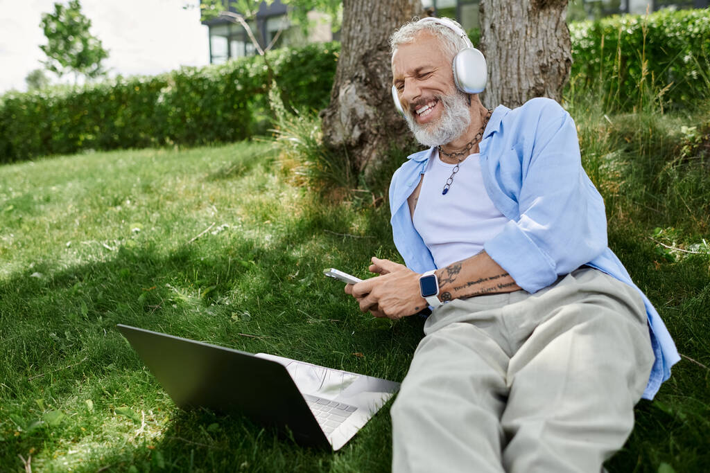 Un hombre maduro con tatuajes y barba gris disfruta de un día al aire libre, escuchando música con auriculares y trabajando en su portátil. - Foto, imagen