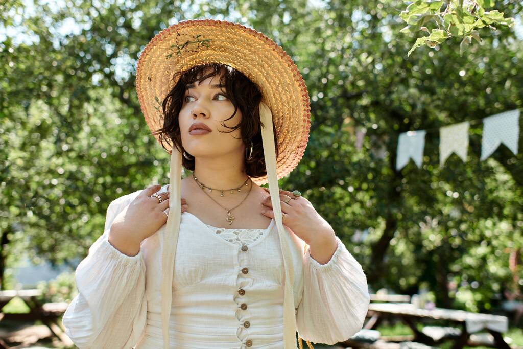 Młoda kobieta w białej bluzce i słomianym kapeluszu patrzy troskliwie w dal, ciesząc się letnim dniem w bujnym ogrodzie. - Zdjęcie, obraz