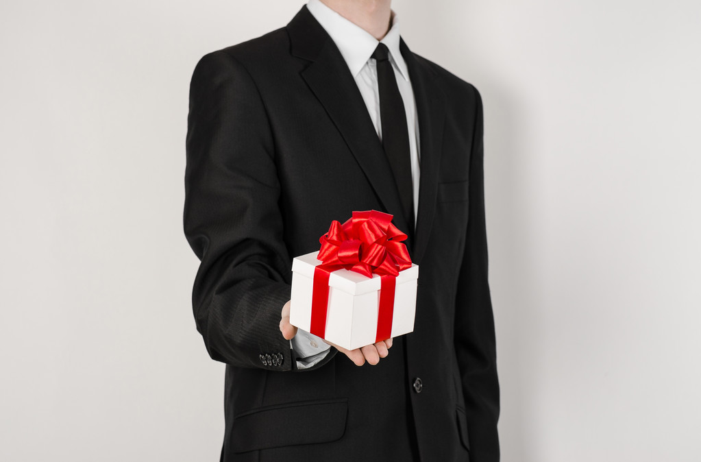 Theme holidays and gifts: a man in a black suit holds an exclusive gift in a white box wrapped with red ribbon and bow isolated on a white background in studio - Fotografie, Obrázek
