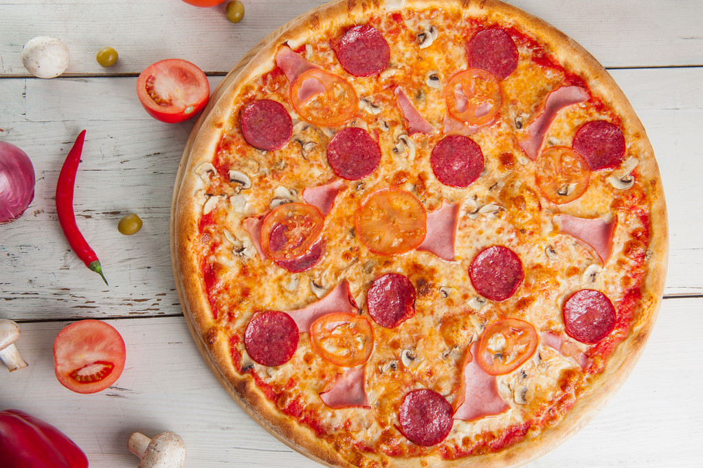 Lähikuva Herkullinen Meat Pizza salami, pekoni ja tomaatti viipaleet leikkuulaudalla valkoisella puupohjalla eri värikkäitä vihanneksia ylhäältä
 - Valokuva, kuva