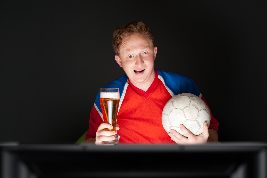 Jovem segurando bola de futebol e cerveja e assistindo TV Translati
 - Foto, Imagem