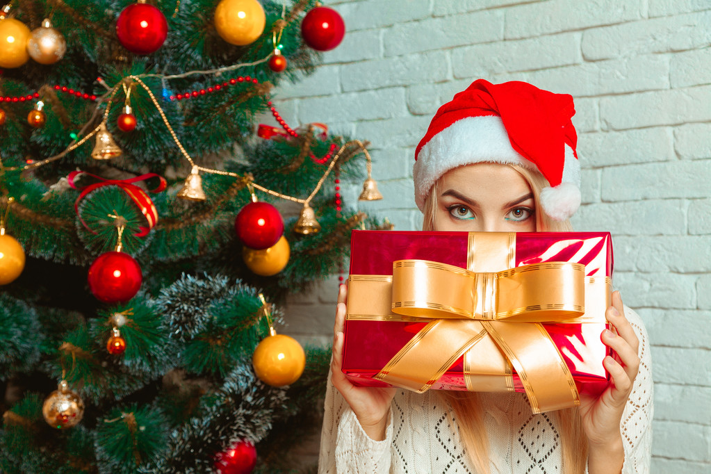 Loira encantadora em Papai Noel esconde seu rosto atrás de um grande presente de Natal
 - Foto, Imagem