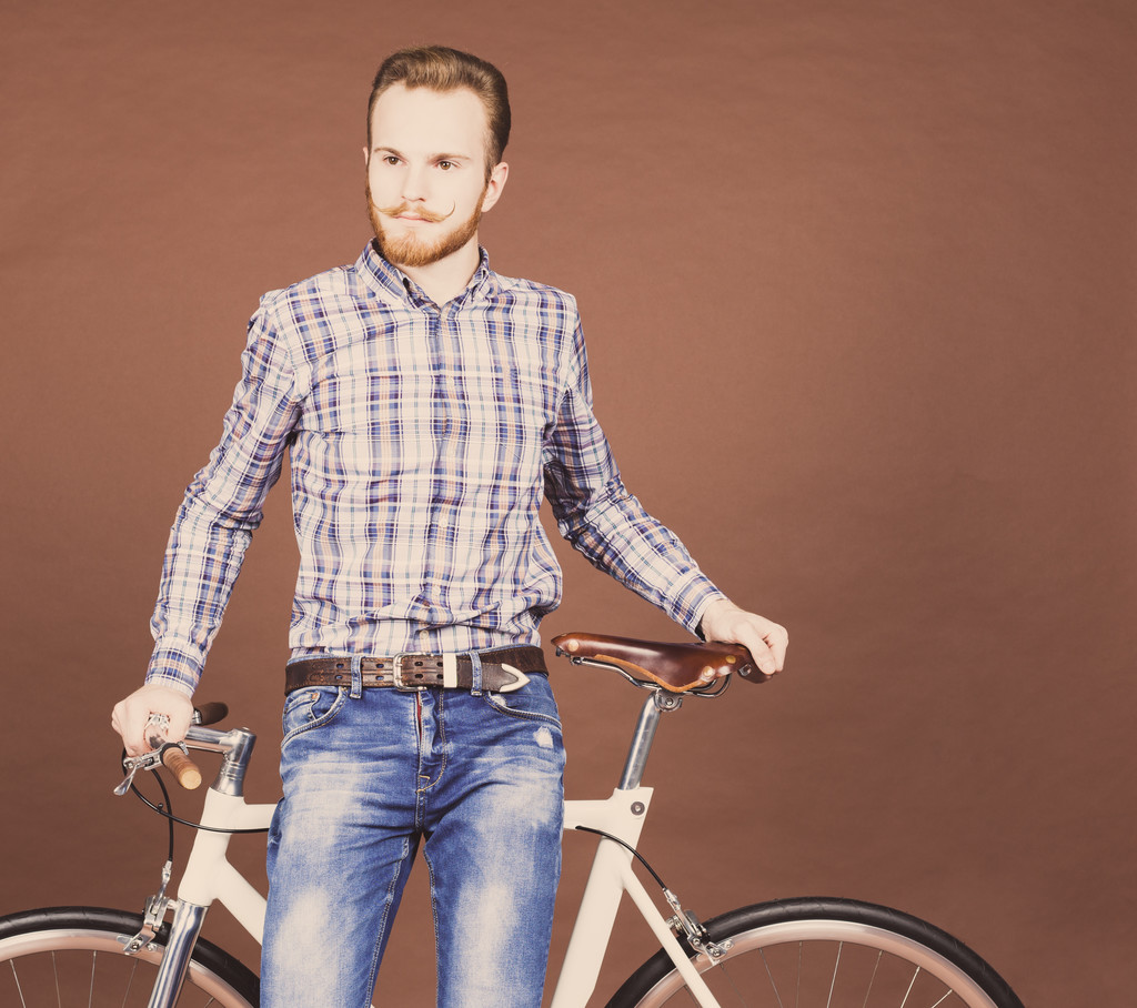 Um jovem com bigode e barba está perto da moderna e elegante bicicleta fixgear. Jeans e camisa xadrez estilo hipster. Cor tonificada
 - Foto, Imagem