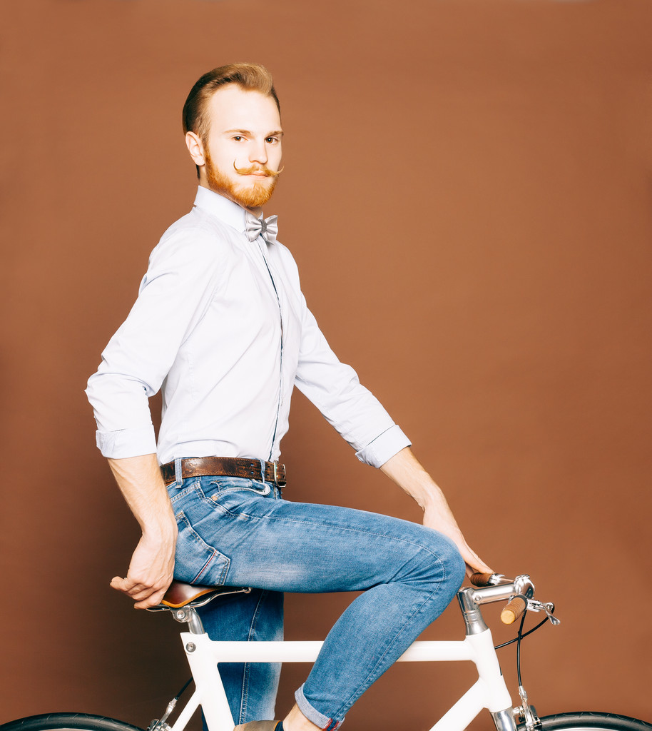 A young man with mustache and beard is sitting on a fashionable modern fixgear bicycle. Jeans and shirt, the bow tie hipster style. Toned color - Photo, Image