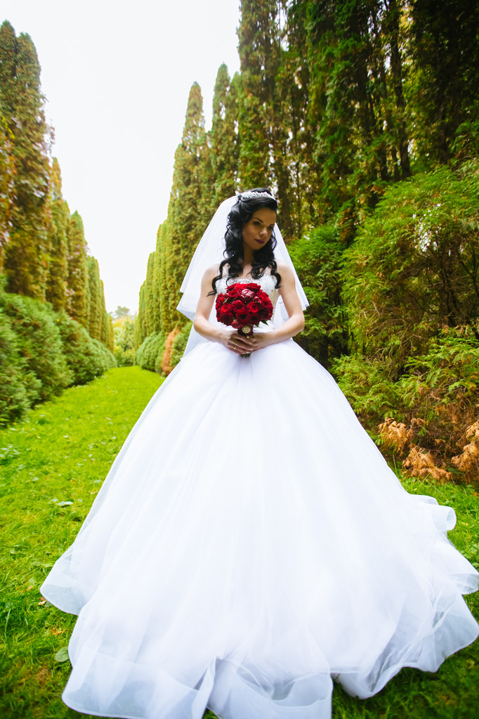 hermoso ramo de color burdeos en las manos de la novia en un vestido blanco de pie en el parque
 - Foto, Imagen