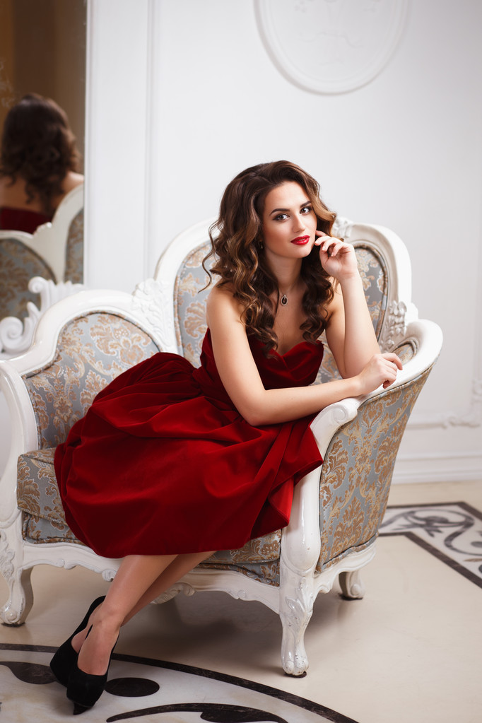 Mooie jonge vrouw met perfecte make-up en haarstijl in prachtige rode avondjurk in dure luxe interieur - Foto, afbeelding