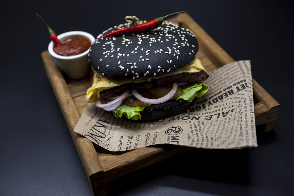 Black burger. A burger with a black roll slices of juicy marble beef, fused cheese, fresh salad and sauce of a barbecue. A burger on the newspaper on a wooden tray  on a dark background. - 写真・画像