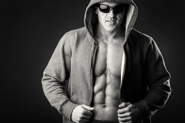 Hombre musculoso con chaqueta deportiva y gafas oscuras sobre fondo negro
. - Foto, imagen