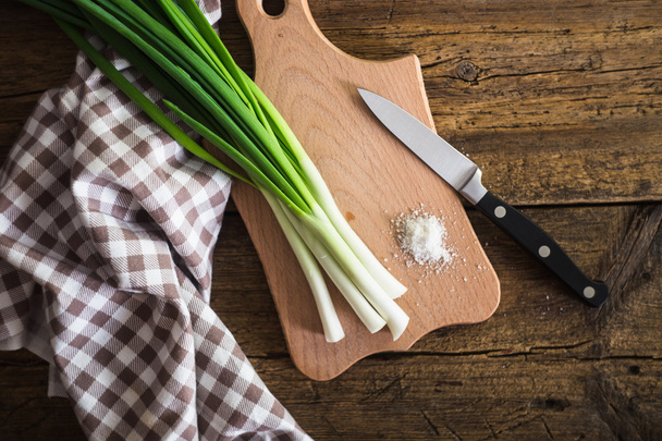 Green onions on a chopping board. Knife and napkin on a wooden table - Photo, Image