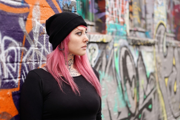 Nainen vaaleanpunainen hiukset lävistykset ja tatuoinnit nojaten graffiti seinään
 - Valokuva, kuva