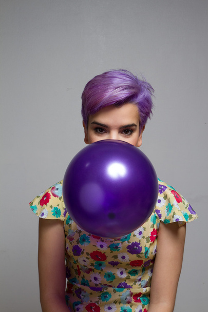 Violeta chica de pelo corto sosteniendo un globo con su boca, mira
 - Foto, Imagen