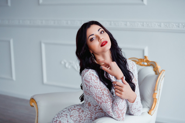 Portrait of perfect, sexy young woman with long black hair and red lips wearing seductive white dress sitting on luxury vintage chair. - Fotoğraf, Görsel